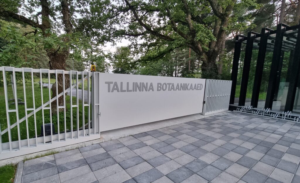 Sissepääsuviit Tallinna Botaanikaaeda aiavärava ja klaashoonega, mida ümbritsevad puud.