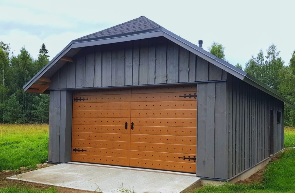 Väike ühekorruseline hall puithoone järsu tumeda katusega ja suure kahekordse puidust garaažiuksega. Struktuur asub rohusel alal, mida ümbritsevad puud ja taustal on pilvine taevas.