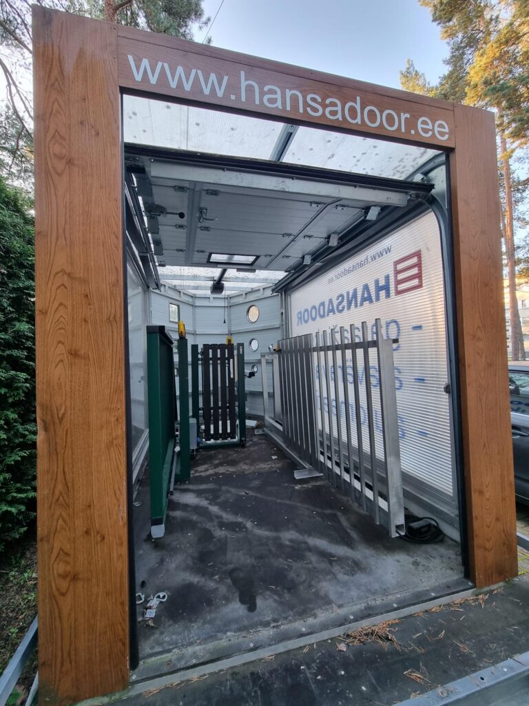 Osaliselt avatud automaatse uksega näidisgaraaž, kus on HANSADOORi reklaambännerid, metallväravad ja leiunurk, mis tutvustab kasutatud garaažiukside valikuid.